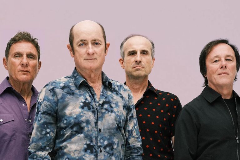 Australian rock band Hoodoo Gurus members standing side-by-side posing in patterned shirts against a light lilac background.